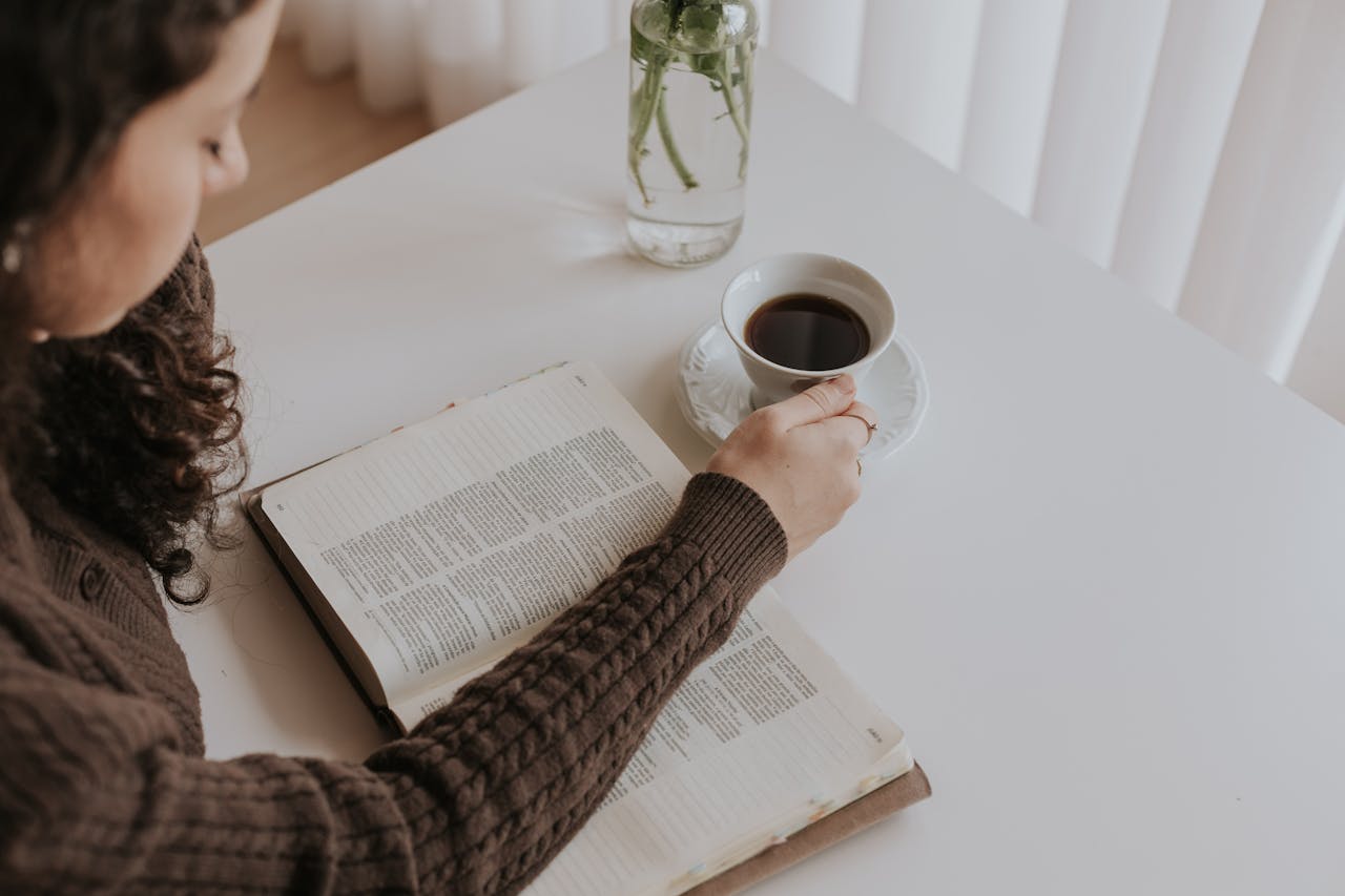 Home A woman enjoys a peaceful moment reading a book and sipping coffee at a bright indoor space.