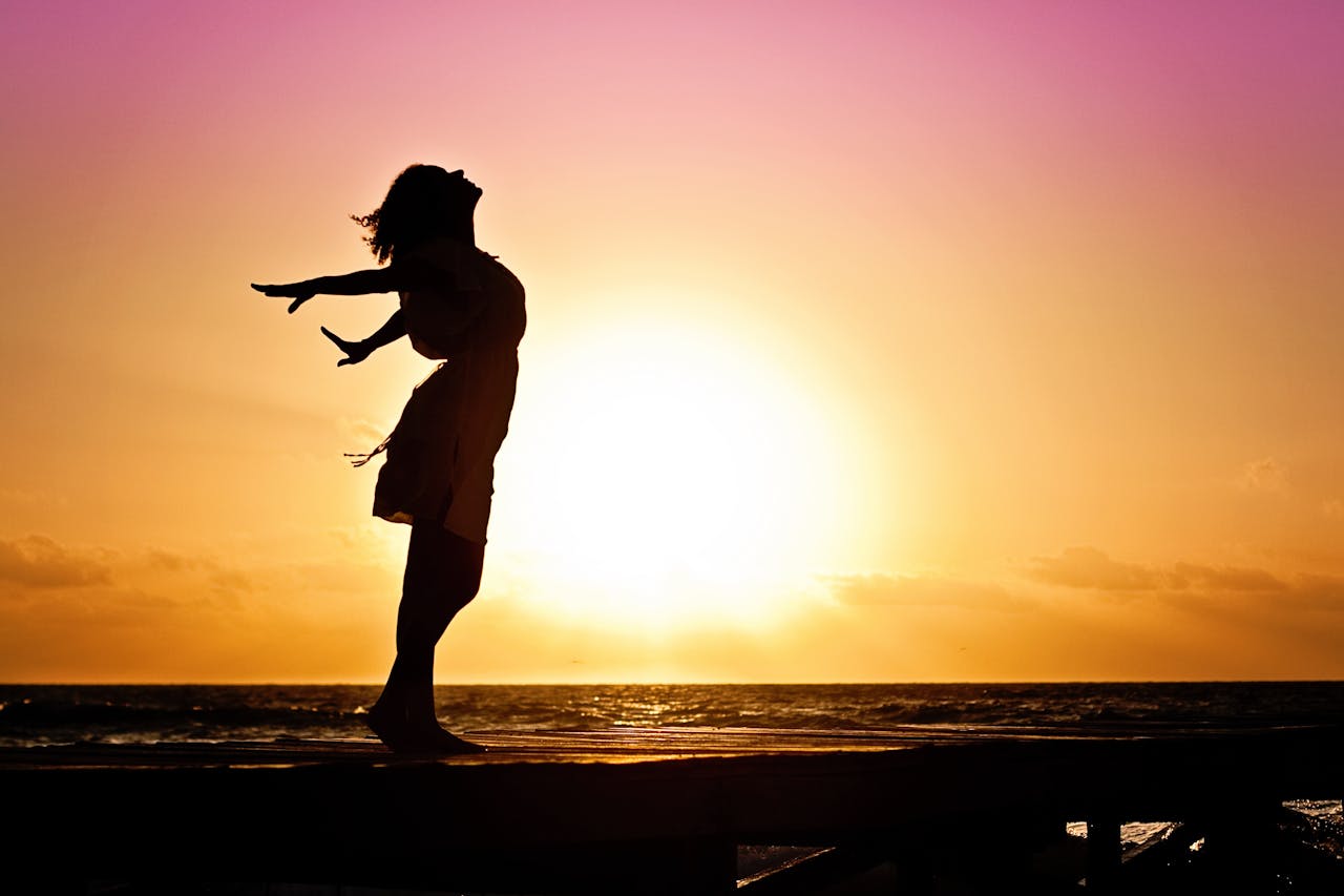 Home Silhouette of a woman at the beach with arms outstretched against a vibrant sunset backdrop.